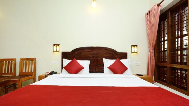 Habitación de hotel con una cama grande, cabecera de madera, dos lámparas de noche, una colcha roja y una ventana con cortinas.
