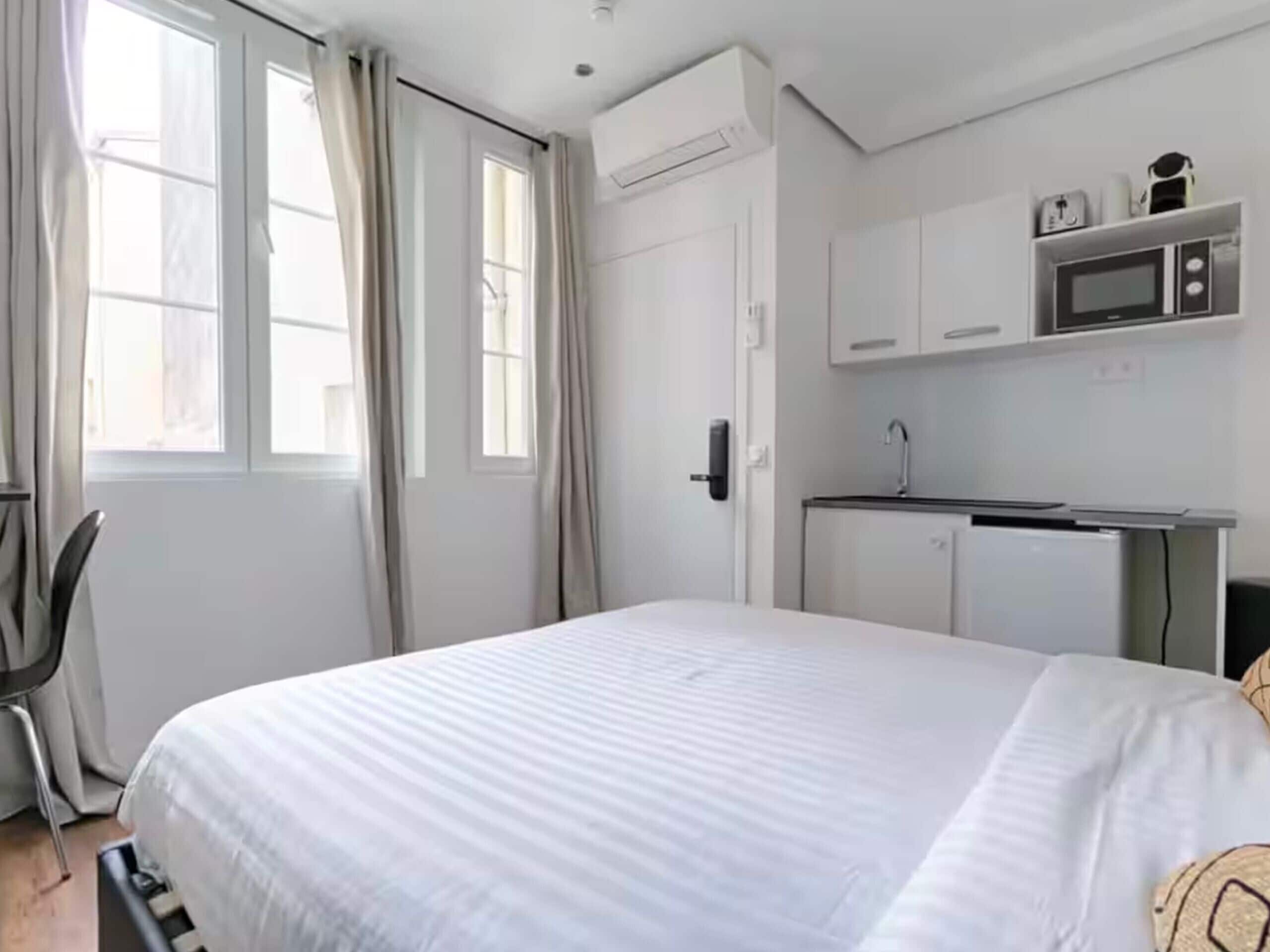Un dormitorio con cama, una puerta, una ventana con cortinas y una kitchenette con microondas, además de un armario pequeño.