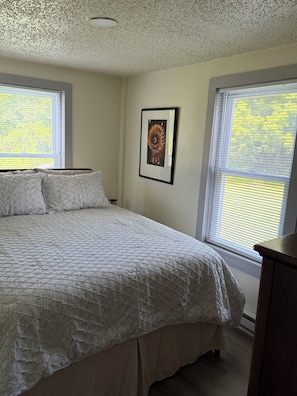 A bedroom with a large bed, two windows with blinds, a framed picture on the wall, and a ceiling vent.