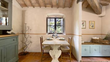 A dining area with a table set for two, a window with a view of greenery, and a cabinet with glassware.