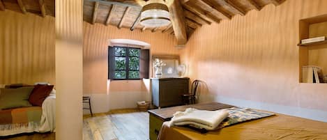 A rustic room with a wooden ceiling, a bed with a white blanket, a wooden desk, and a window with a view of greenery.