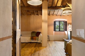A room with a wooden floor, a bed with a blanket, a window with a view, and a hanging light fixture.