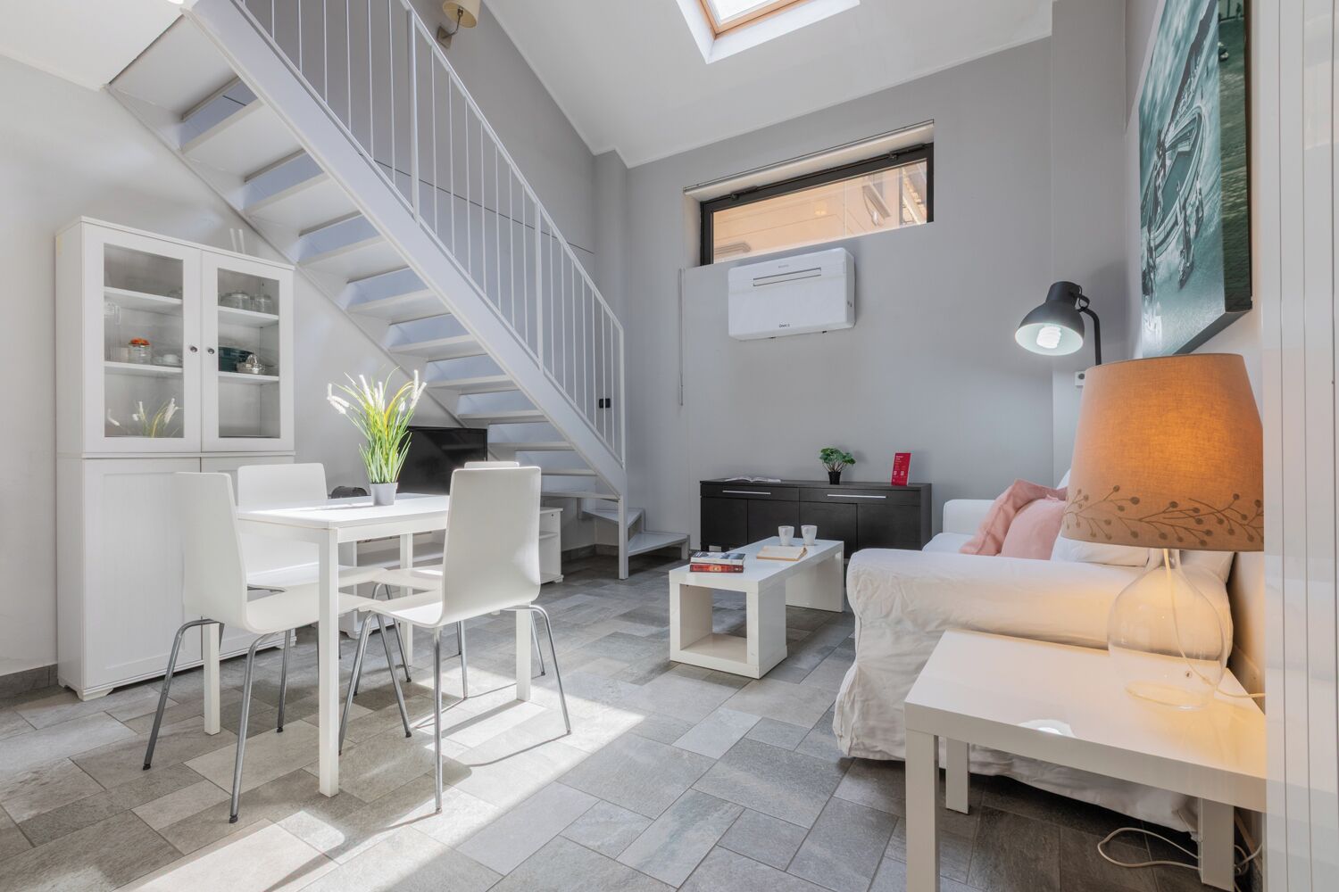 A modern living space with a staircase, white furniture, a dining area, and a sofa with a lamp.