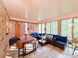 A sunroom with a wooden table, blue sofa, and large windows offering a view of trees.