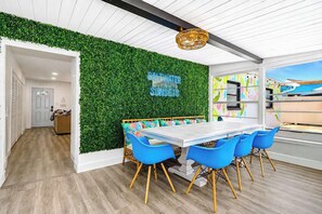 A room with a white ceiling, a green wall with a neon sign, a wooden bench, and a table with blue chairs.