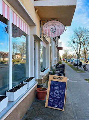 Ein Schaufenster mit einem Schild für das Café 'Eisschleuse 5', Sitzgelegenheiten im Freien mit Blumentöpfen und einer Kreidetafel mit Speisekarte.