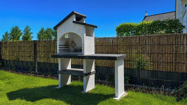 Een barbecue met schoorsteen, voor een bamboe hek en een gazon.