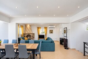 A modern living room with a stone fireplace, a sofa set, a dining table with chairs, and a television. - Villa Starlight - Five Bedroom Villa, Sleeps 11 (Lardos)