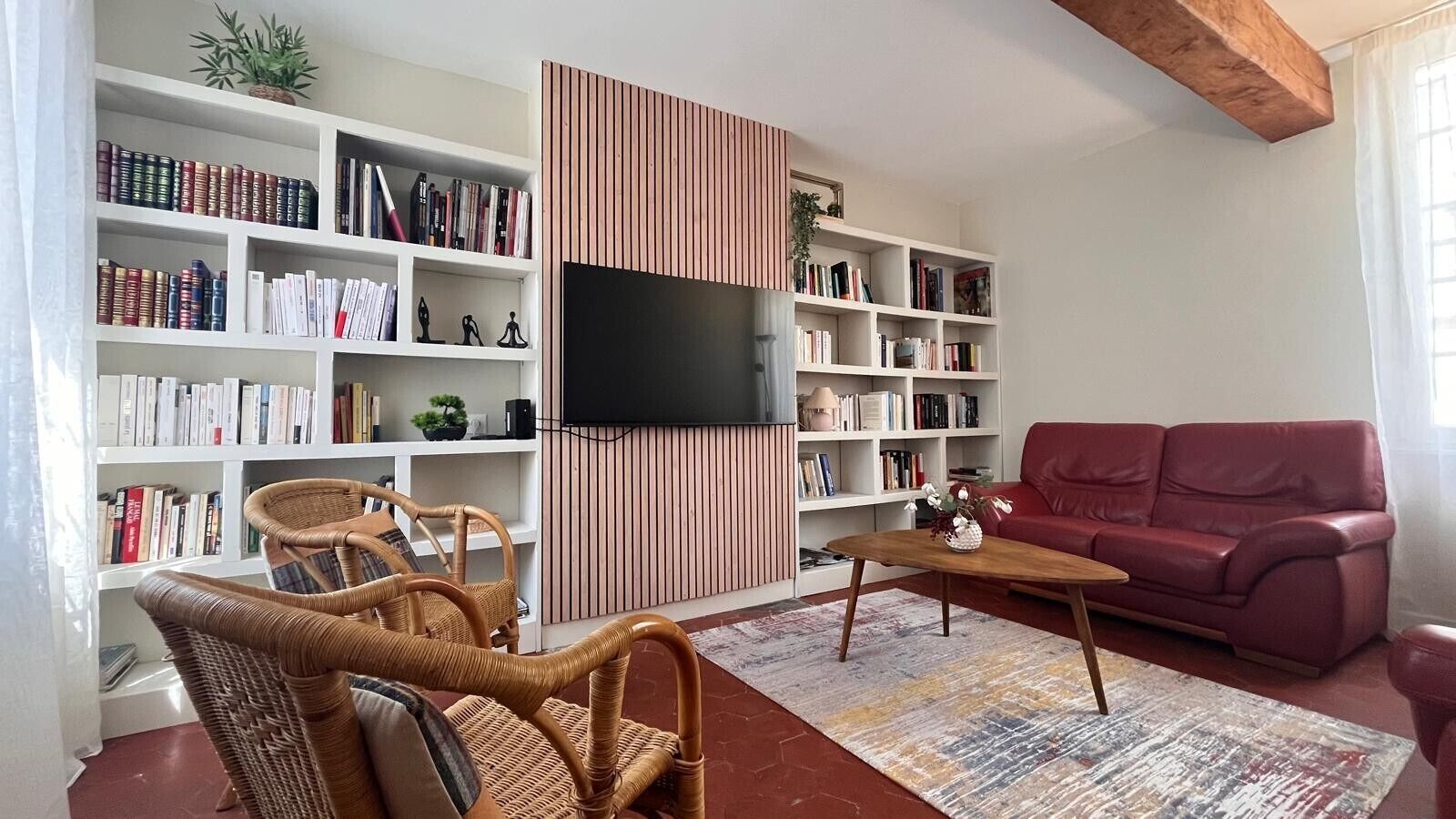 Een woonkamer met een boekenkast, een flatscreen televisie, een rode bank en rieten stoelen.