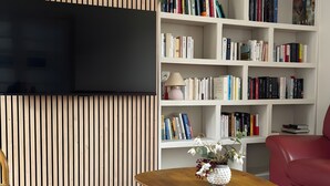 Una sala de estar con un televisor de pantalla plana montado en una pared de madera, una estantería blanca llena de libros, un sofá rojo y una mesa de centro de madera con un jarrón de flores.