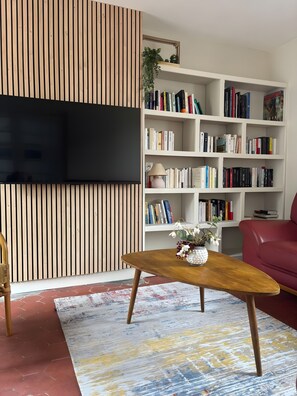 Ein Wohnzimmer mit einem an einer Holzwand montierten Flachbildfernseher, einem weißen Bücherregal voller Bücher, einem roten Sofa und einem Holztisch mit einer Blumenvase.