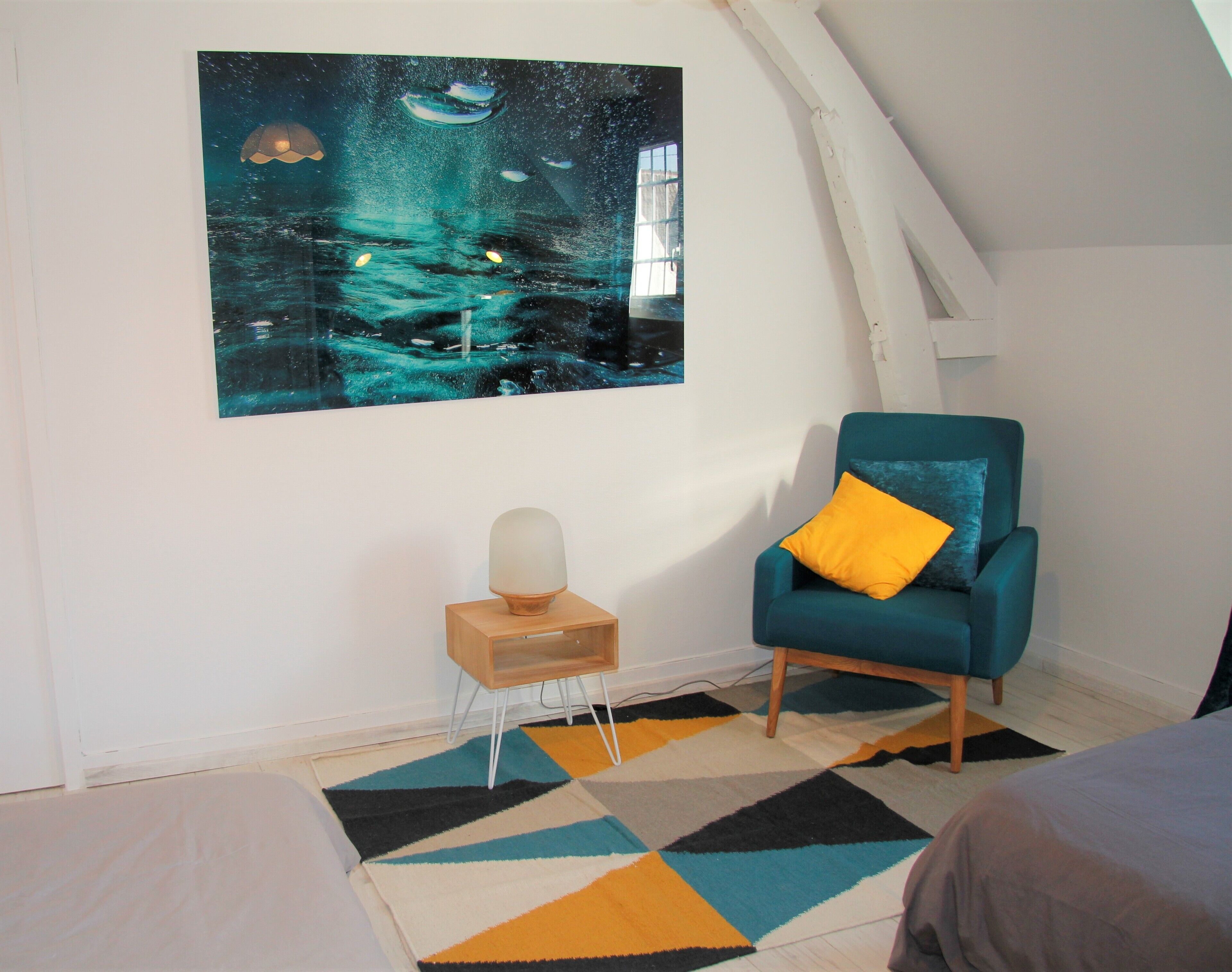 Een kamer met een blauwgroene fauteuil, een klein tafeltje met een lamp, een kleurrijk vloerkleed en een groot abstract schilderij aan de muur.