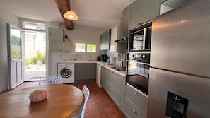 A modern kitchen with a built-in washing machine, a dining area with a wooden table, and a view of the outside through a glass door.