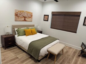 Ein Schlafzimmer mit einem Bett, Nachttischen, einer Bank, einem Fenster mit Jalousien und einem Wandbild.