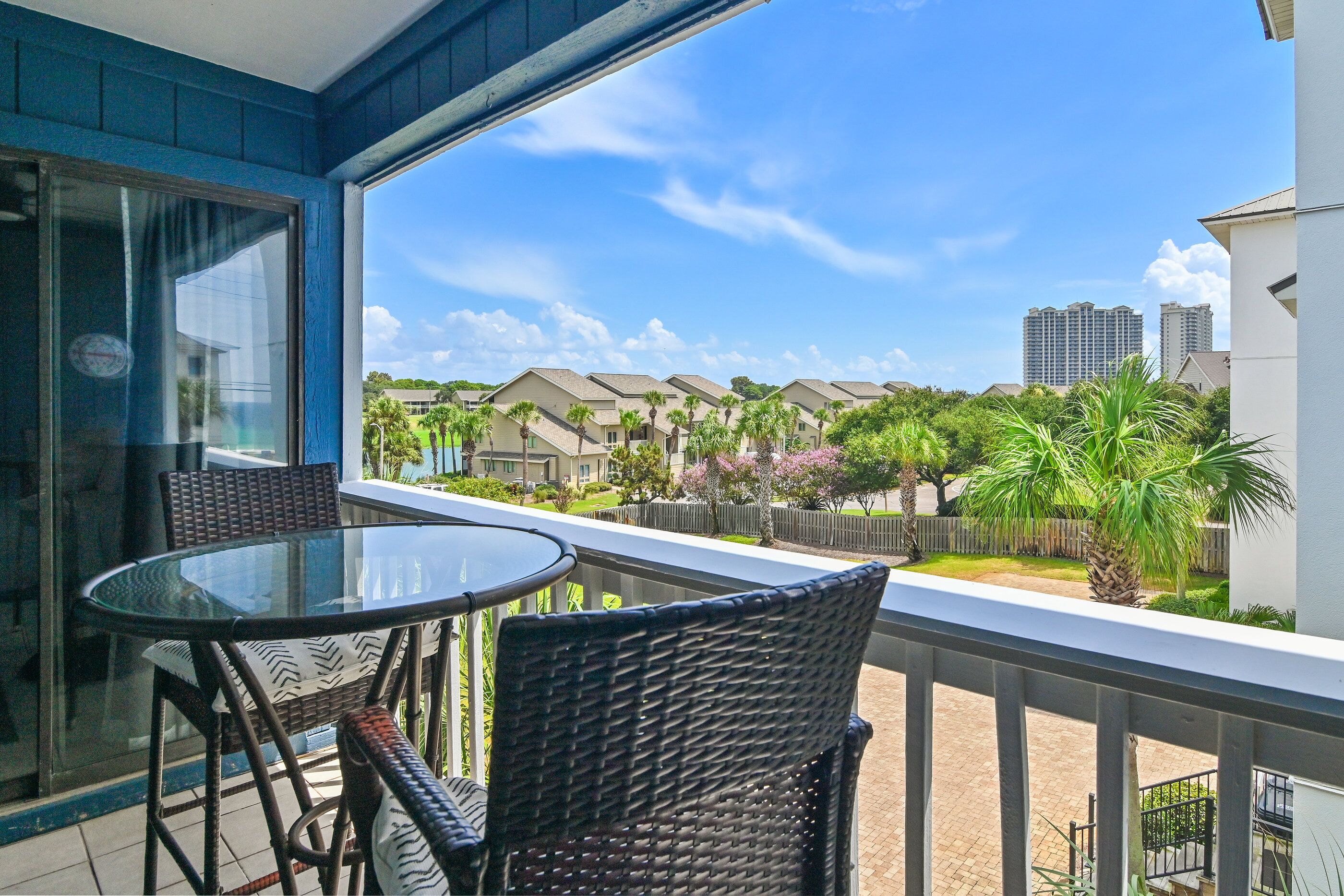 A balcony with two wicker chairs and a glass table, overlooking a residential area with houses and palm trees.