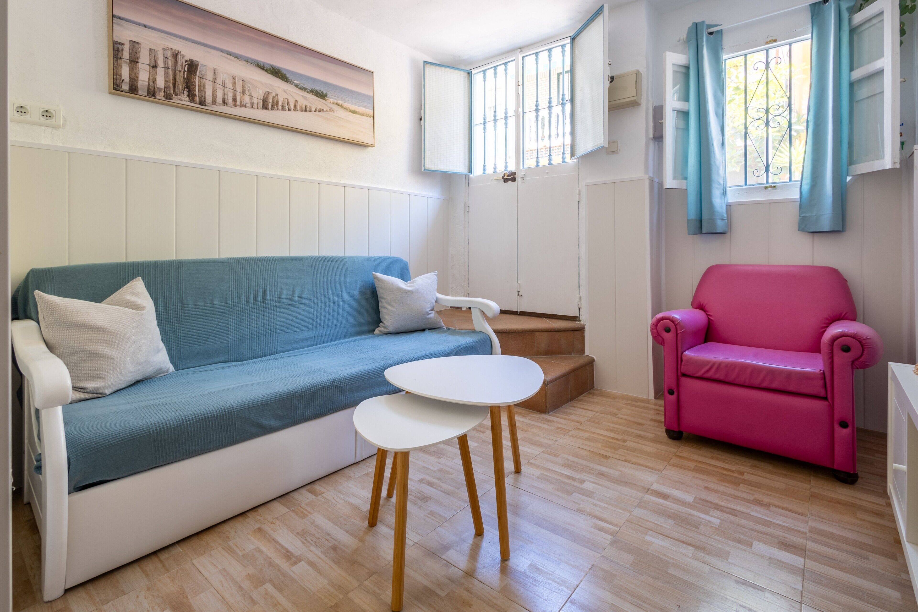 A room with a blue sofa, a pink armchair, a small white table, and a painting on the wall.