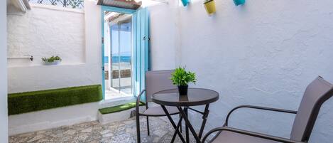 Une terrasse avec une table et des chaises, une petite plante en pot et une vue sur la mer par une porte ouverte.