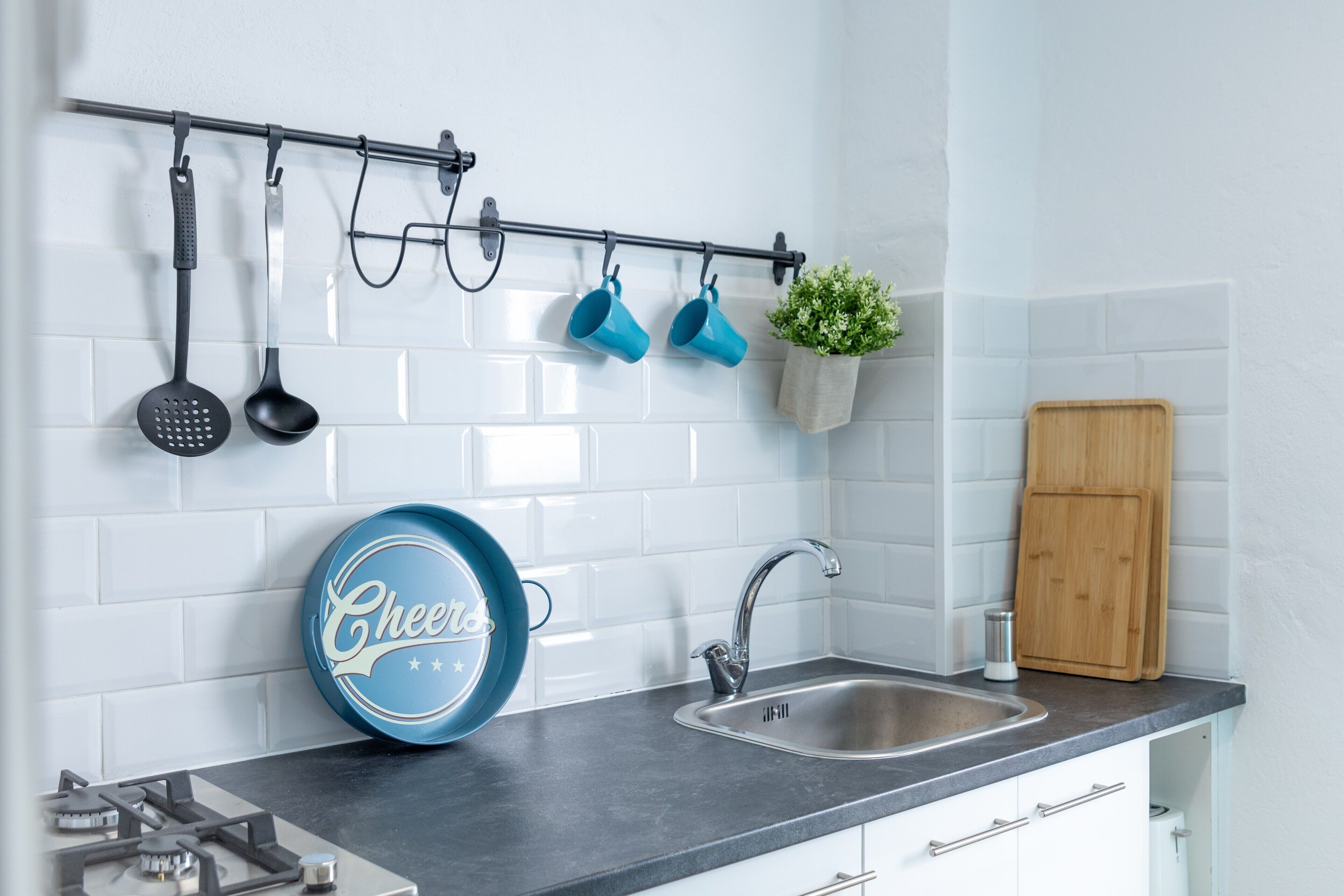 A kitchen with a stainless steel sink, a gas stove, a hanging rack with kitchen utensils, and a blue plate with the word 'Cheers'.