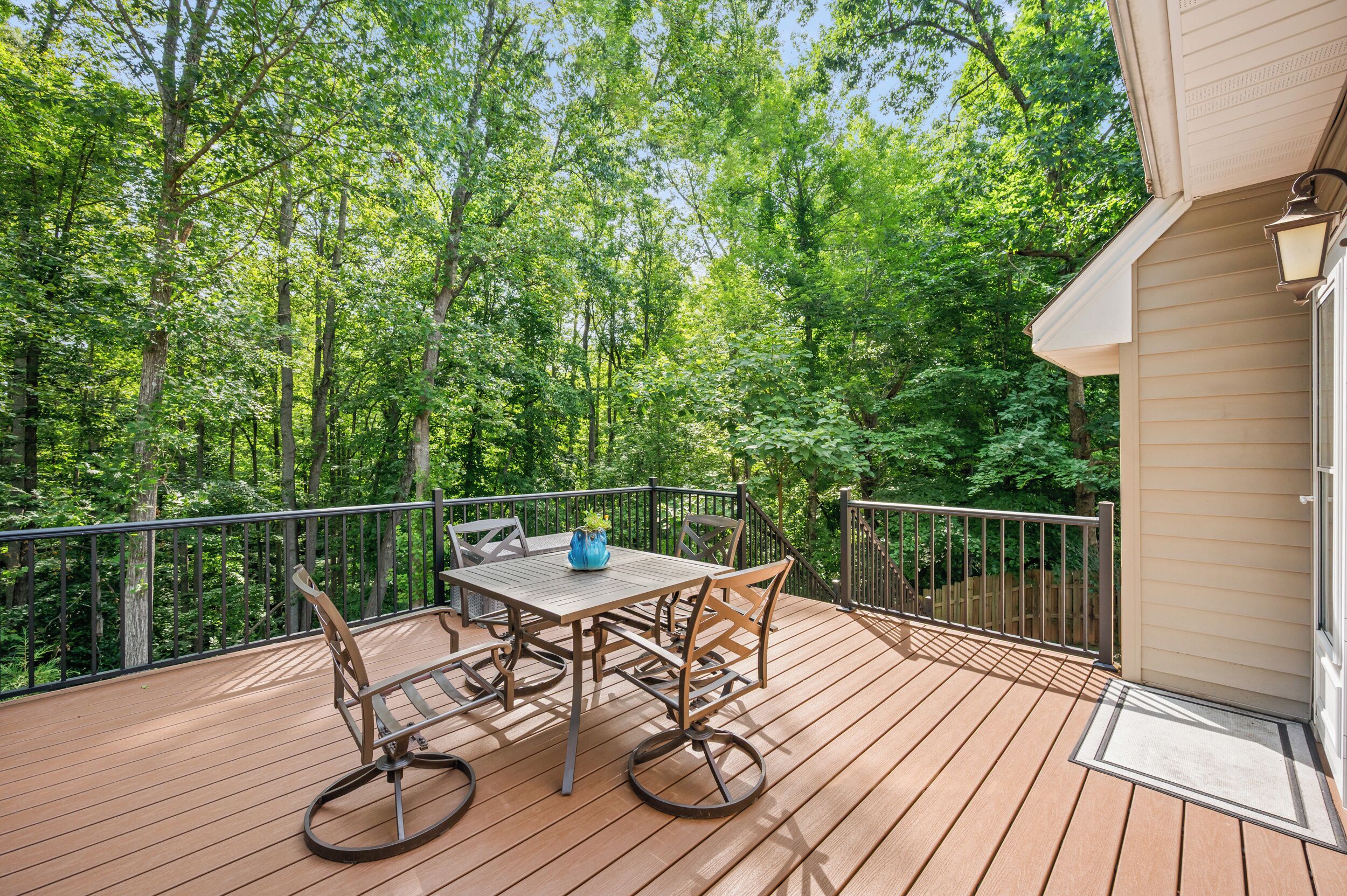 A wooden deck with a table and chairs, surrounded by a metal railing, overlooking a forested area.