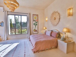 Une chambre avec une grande fenêtre, un lit recouvert d’une couvre-lit rose, deux lampes de chevet et un objet décoratif fixé au mur.