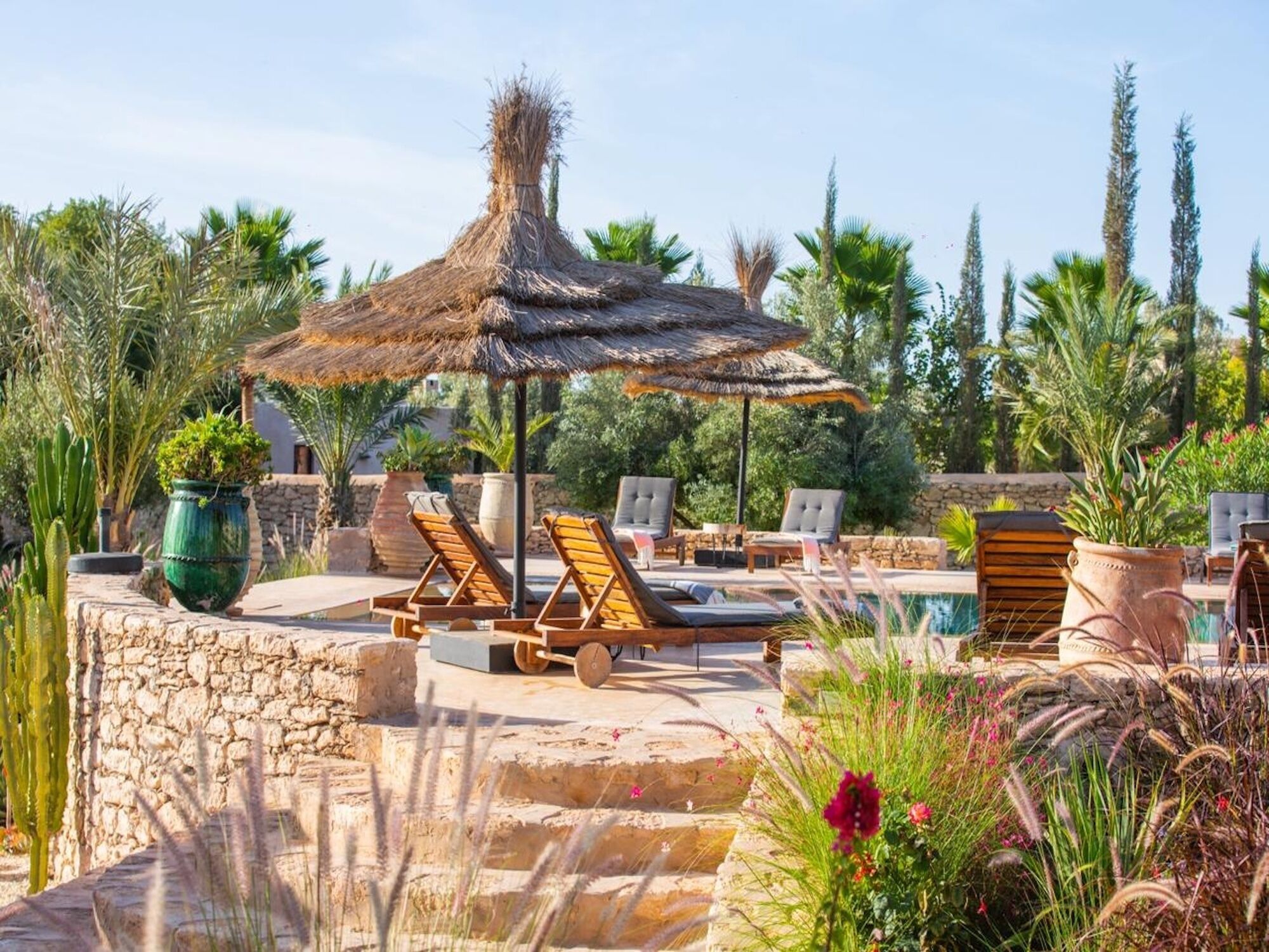 Ein Poolbereich mit Liegestühlen und einem Reedsdach-Sonnenschirm, umgeben von Grünflächen und Steinmauern.