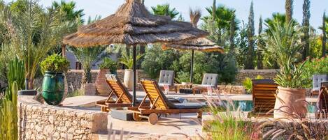 Un espace aménagé au bord de la piscine, avec des chaises longues et un parasol en chaume, entouré de verdure et de murs en pierre.