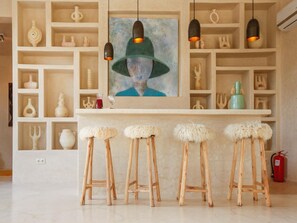 A bar area with wooden stools, a painting above the counter, and shelves with various decorative items. - Palais Baraya (Essaouira)