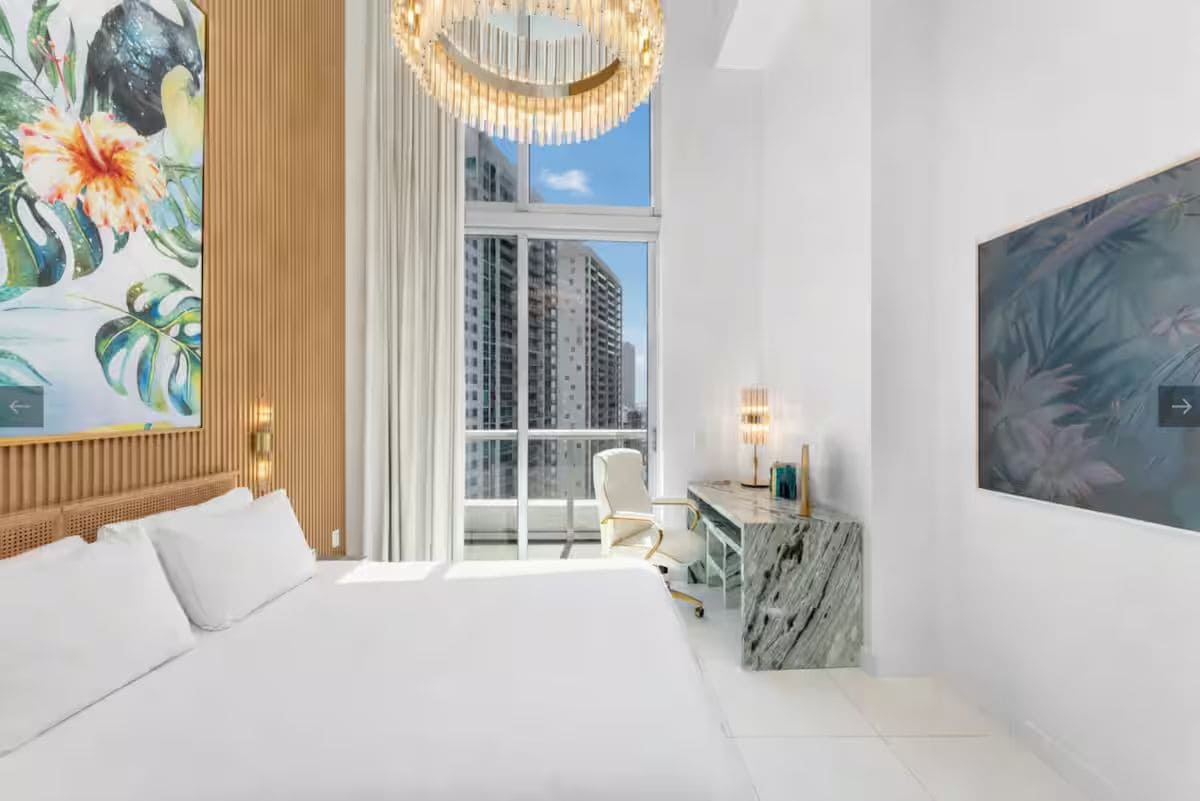 A hotel room with a large bed, a desk with a marble top, a large window with a view of buildings, and a painting on the wall.