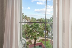 A view through a window with white curtains, featuring a courtyard with palm trees and a red-roofed building. - 1BR apartment with 2 queens near Disney (Orlando)