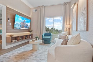A modern living room with a flat-screen TV, a sofa, a coffee table, and a view of palm trees through the window. - 1BR apartment with 2 queens near Disney (Orlando)
