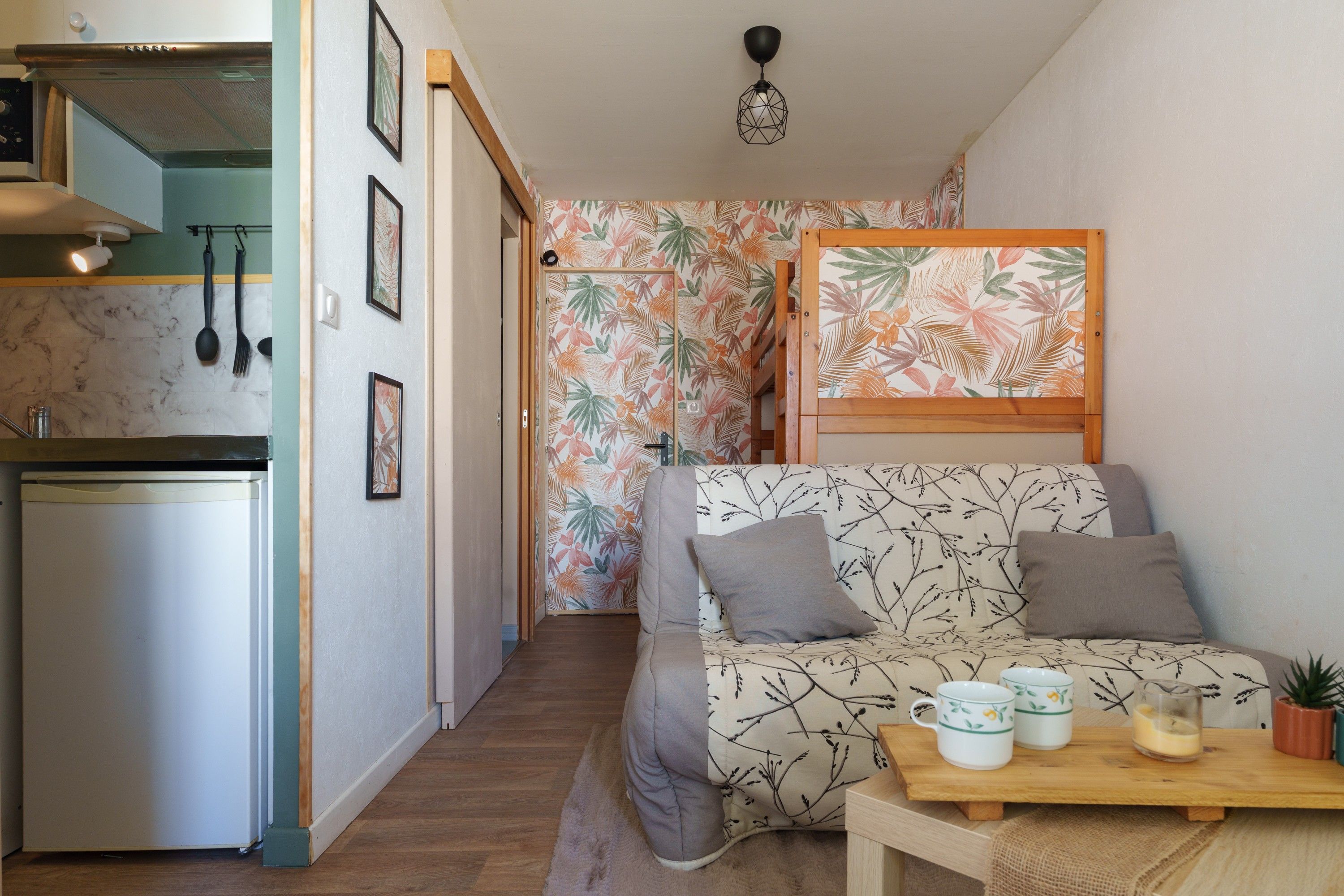 Een knus woongedeelte met een kitchenette, een bank met kussens, een houten tafel met kopjes en een glas, en een kamer met een behang.