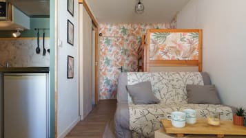Een knus woongedeelte met een kitchenette, een bank met kussens, een houten tafel met kopjes en een glas, en een kamer met een behang.