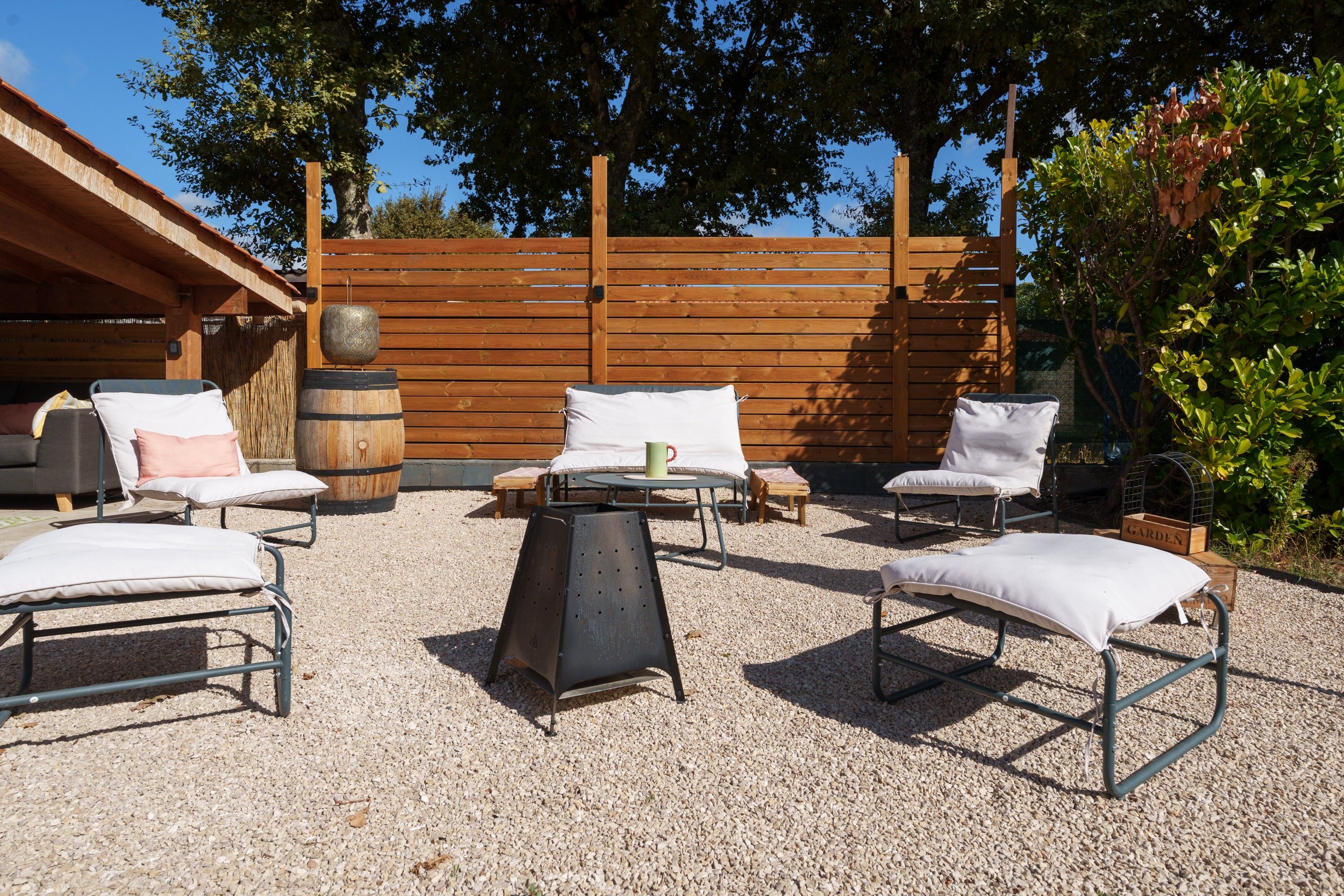 An outdoor seating area with metal chairs and a table, surrounded by a wooden fence and trees.