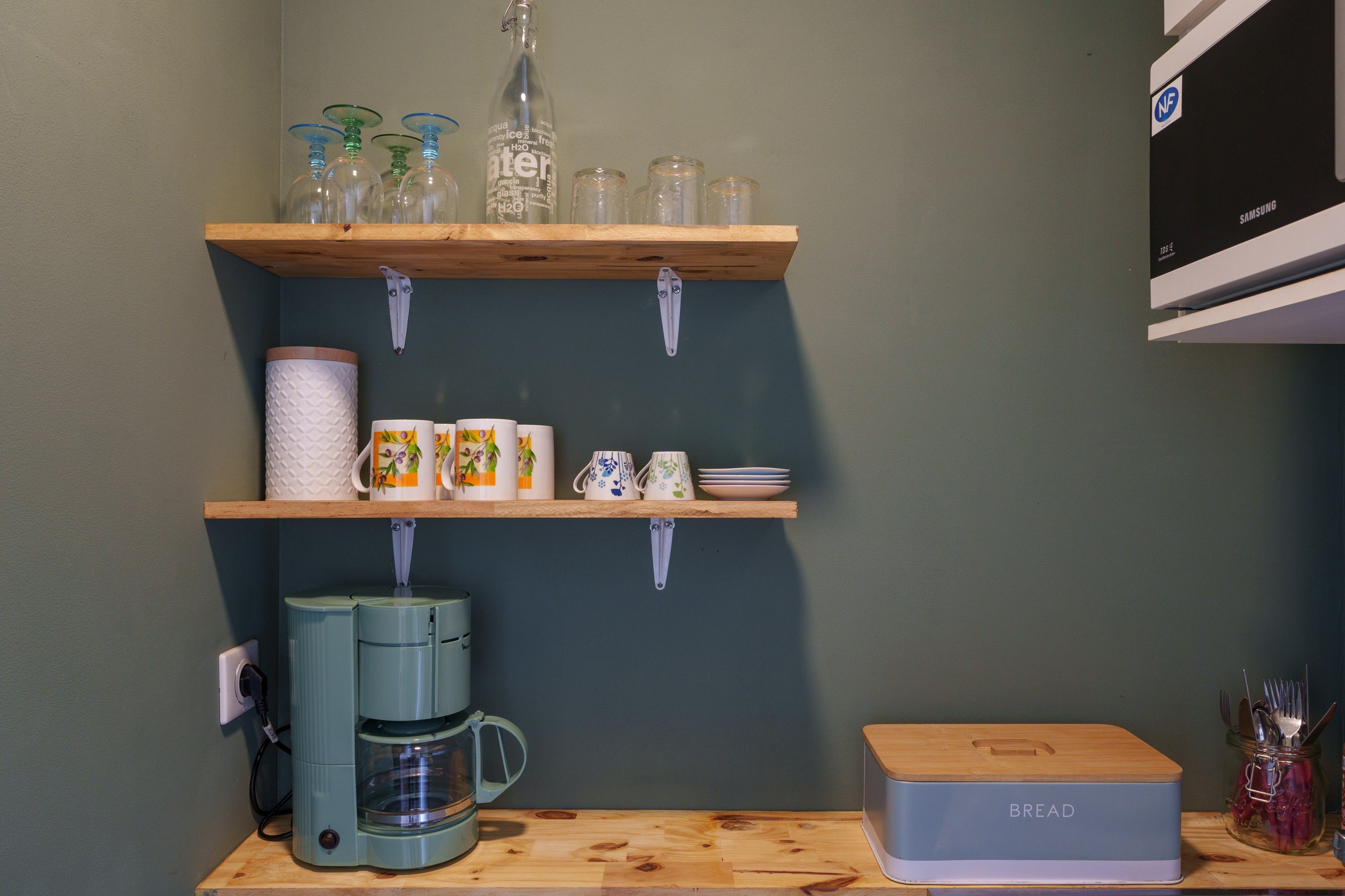 Et køkken med hylder i træ, der indeholder vinglas, krus og en kaffemaskine.