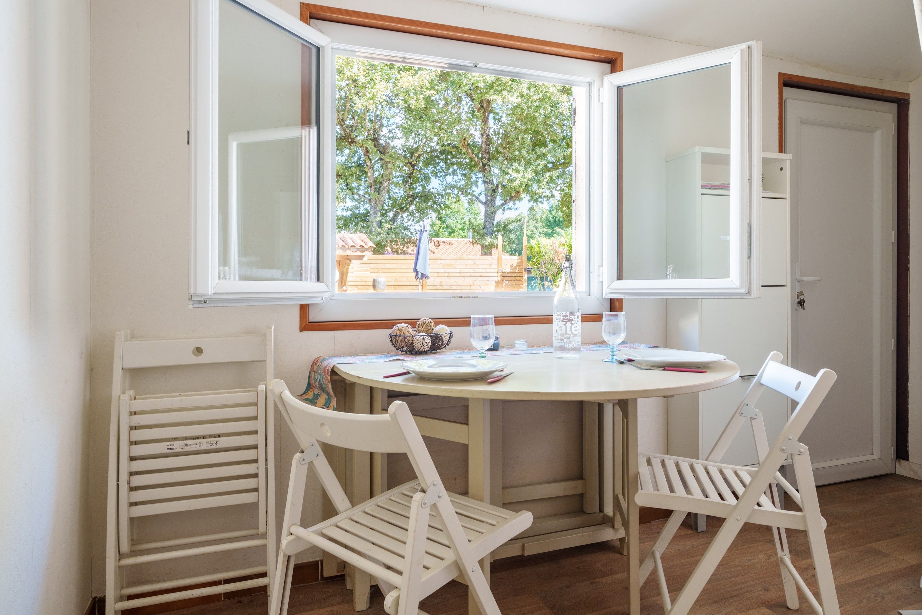 Un comedor con una mesa redonda, dos sillas blancas, una ventana que ofrece vistas abiertas a un jardín y una puerta a la derecha.