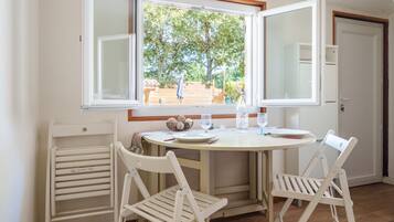 Un comedor con una mesa redonda, dos sillas blancas, una ventana que ofrece vistas abiertas a un jardín y una puerta a la derecha.