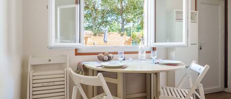 A dining area with a round table, two white chairs, a window with an open view of a garden, and a door to the right.