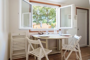 A dining area with a round table, two white chairs, a window with an open view of a garden, and a door to the right.