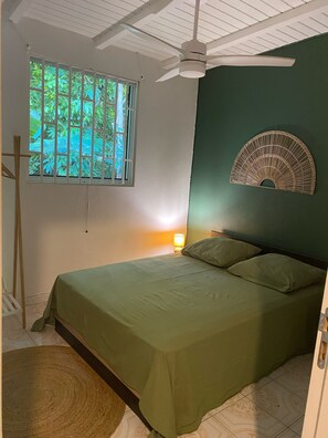 A room with a bed, a ceiling fan, a window with bars, a wall-mounted decorative item, and a bedside lamp. - Creole House on the Beach - Double Bed - Free Parking (Deshaies)