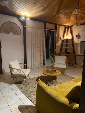 Ein Zimmer mit hohen Decken, einer Holzbalkendecke, einer weißen Tür, einem weißen Stuhl, einem gelben Sofa, einem runden Couchtisch und einem an der Wand befestigten Regal.