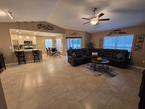 A spacious living area with a black leather sofa, a dining table with chairs, a kitchen area, and a ceiling fan.