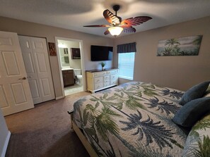 A bedroom with a bed, a ceiling fan, a dresser, a TV, and a painting on the wall. - Terrific Pool Home, close to Flagler Beach & Daytona (Palm Coast)