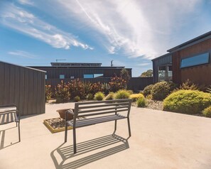 A modern outdoor seating area with a bench and a table, surrounded by a garden with various plants and a building in the background. - Villa Callista - Modern Coastal Escape (Port Sorell)