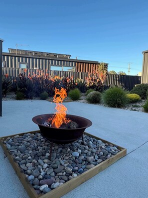 A fire pit with flames, surrounded by a gravel bed, in a landscaped outdoor area with a wooden fence and various plants. - Villa Callista - Modern Coastal Escape (Port Sorell)