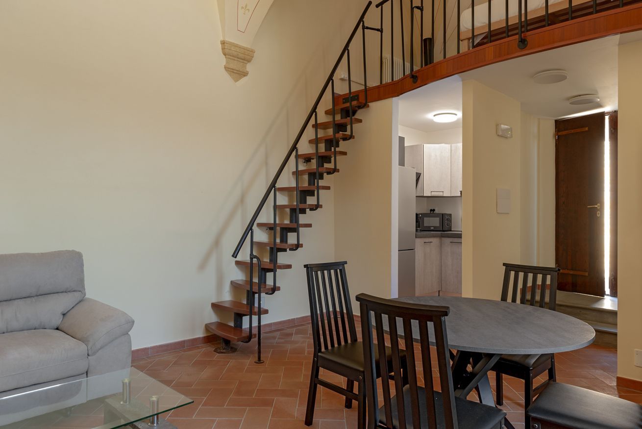 A living room with a sofa, a glass coffee table, a dining table with chairs, and a staircase with wooden steps and black railings.