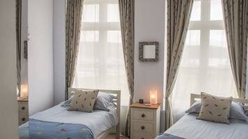A bedroom with two single beds, a bedside table with a lamp, a mirror on the wall, and a window with curtains.