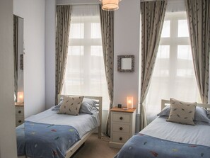 A bedroom with two single beds, a bedside table with a lamp, a mirror on the wall, and a window with curtains. - 4 The Salcombe (Salcombe)