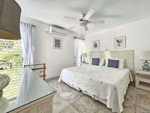 A bedroom with a bed, a ceiling fan, a window with curtains, and a bedside table with a lamp. - Poolside condo with garden access direct to beach! (Holetown)