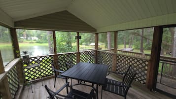 A covered outdoor deck with a table and chairs, overlooking a body of water and a grassy area.