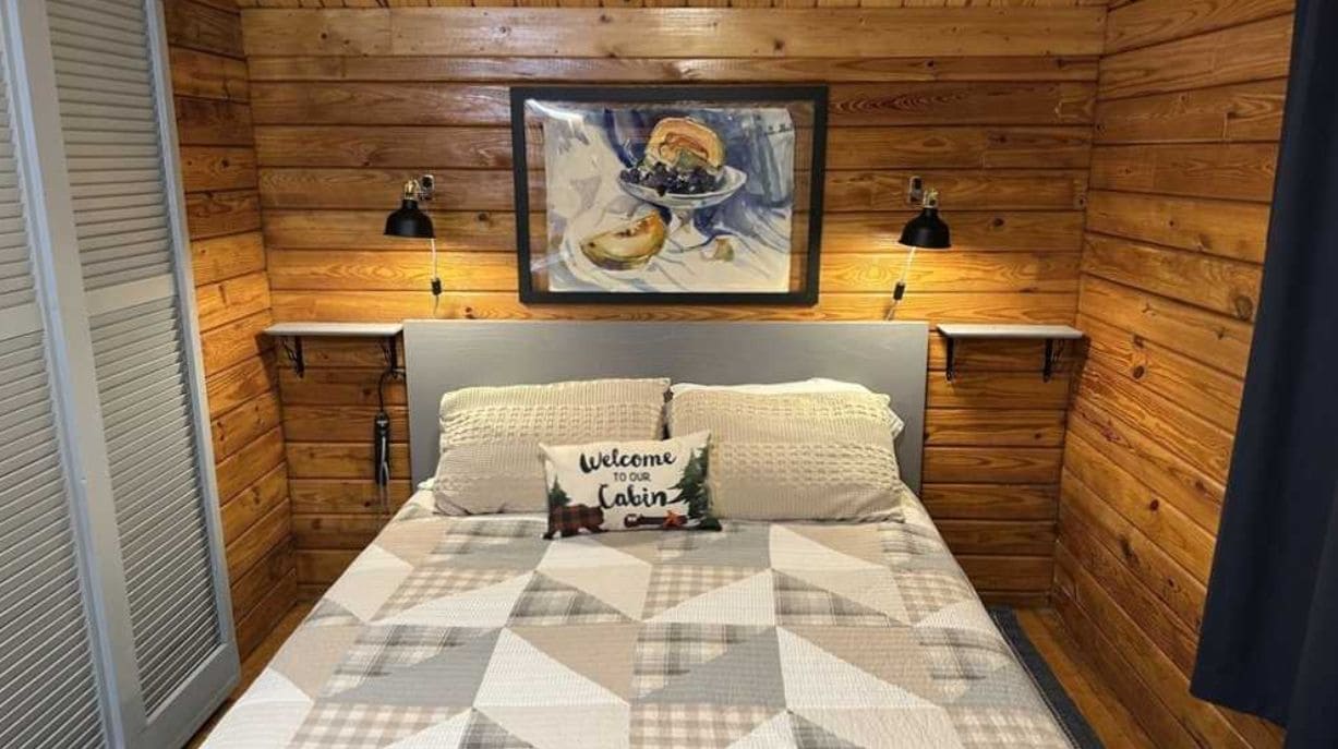 A cabin-style bedroom with a bed, a welcome sign, a framed picture, and two wall-mounted lamps.
